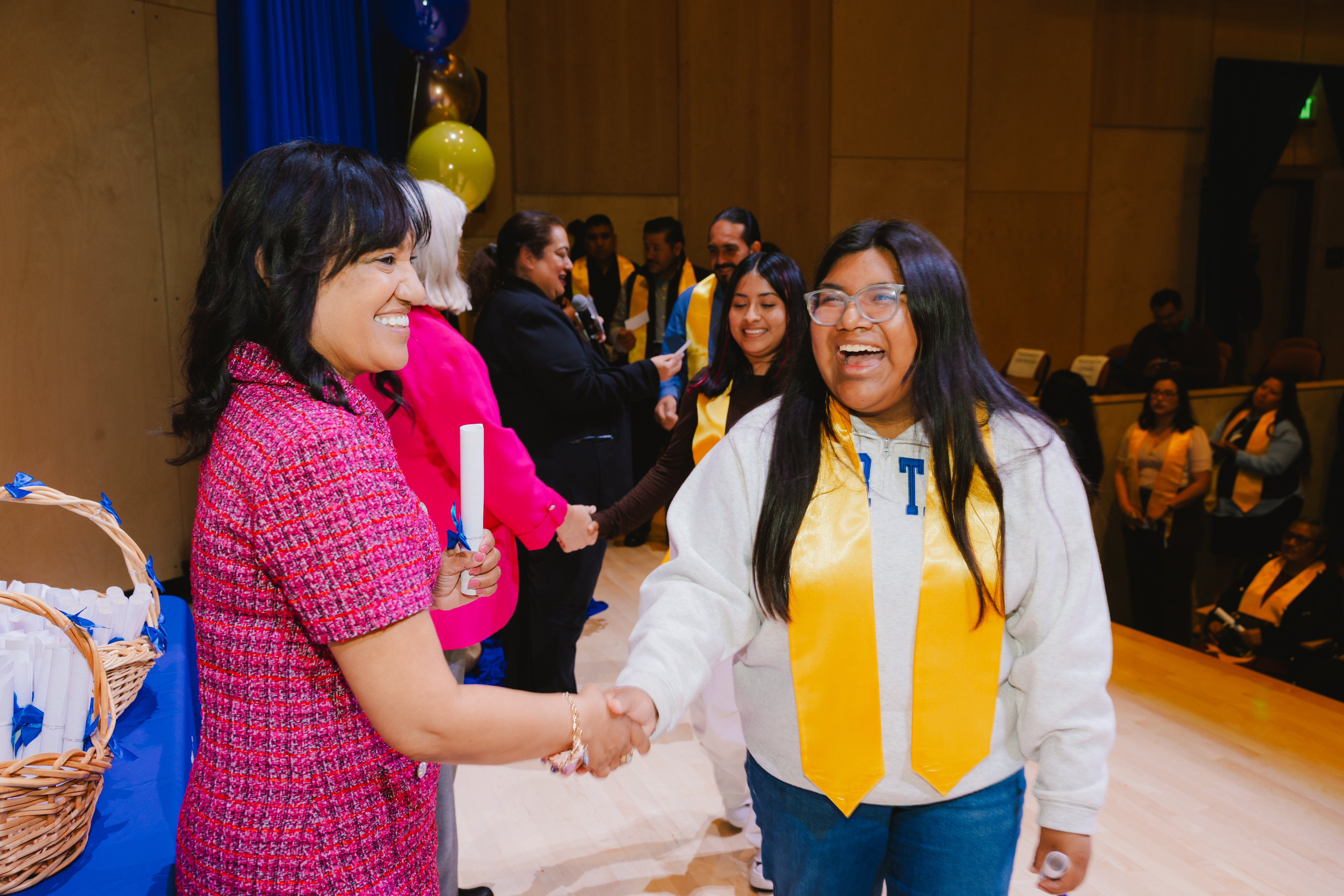A student wearing a yellow stole smiles while shaking hands with a presenter on stage during Allan Hancock College’s Noncredit Recognition Ceremony at Boyd Concert Hall. Other students wait in line behind them as certificates are presented, celebrating completion of noncredit programs and educational milestones.